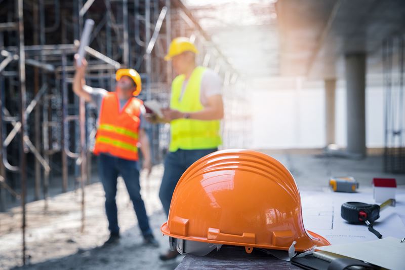 A close-up view of an orange safety hardhat. In the background there are two construction workers in orange vests. The Background is blurry.